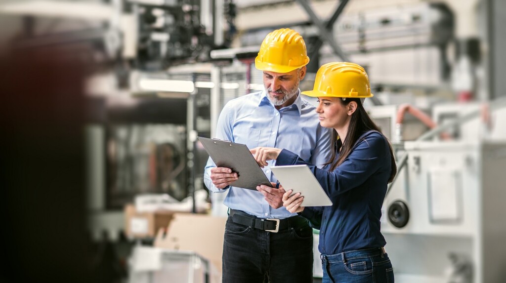 Due professionisti in ambito industriale, dotati di dispositivi di protezione individuale, consultano documentazione tecnica su tablet durante un controllo in fabbrica.